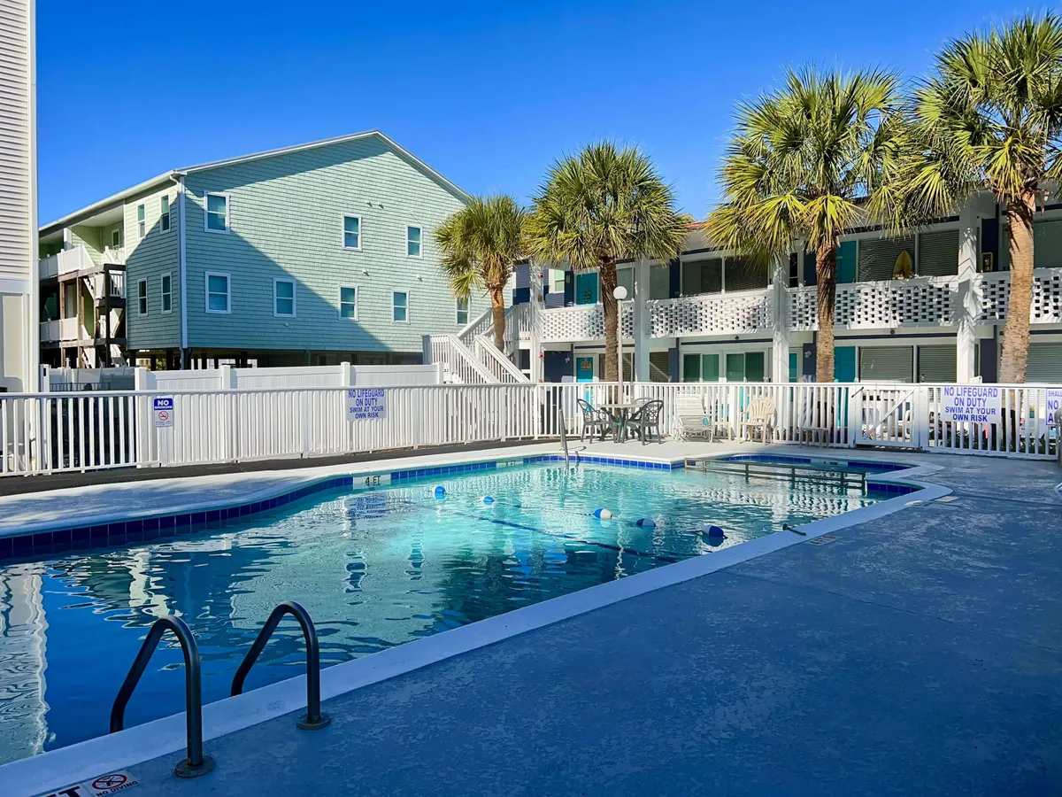 The swimming pool area is surrounded by palm trees, a green building, and comfy outdoor seating. The setting feels relaxed and sociable, with space for swimming, sunbathing, and group gatherings after a day at the beach.