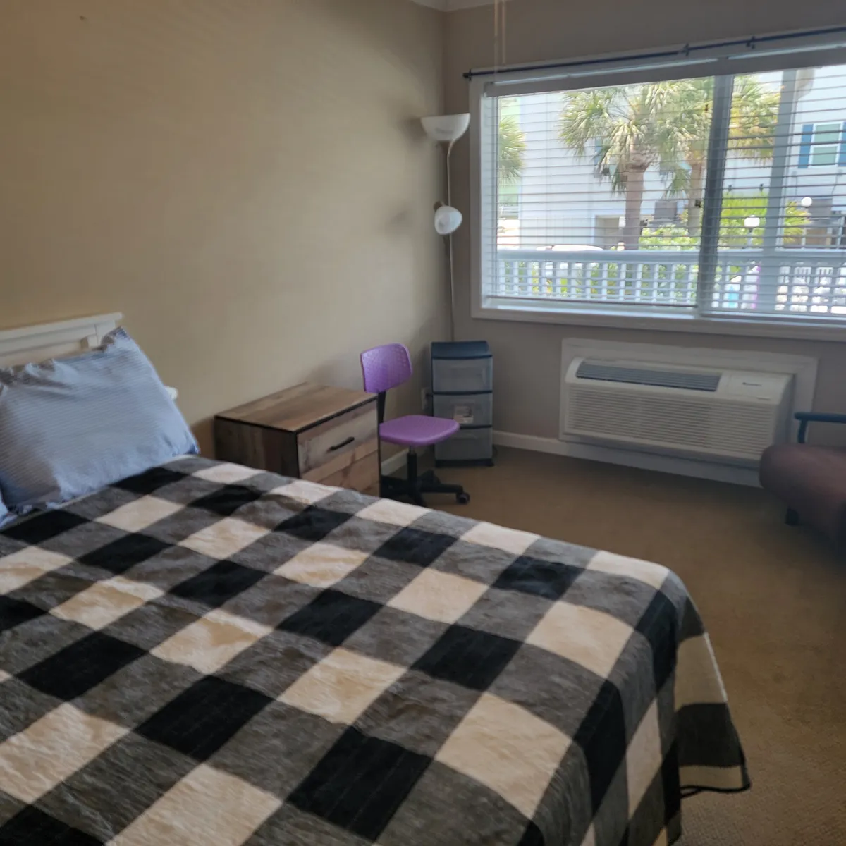 A comfortable bedroom showcasing a bed with a plaid blanket near a sunlit window, accompanied by a small desk and chair. The space appears bright and casual, highlighting both restful and functional elements for guests.