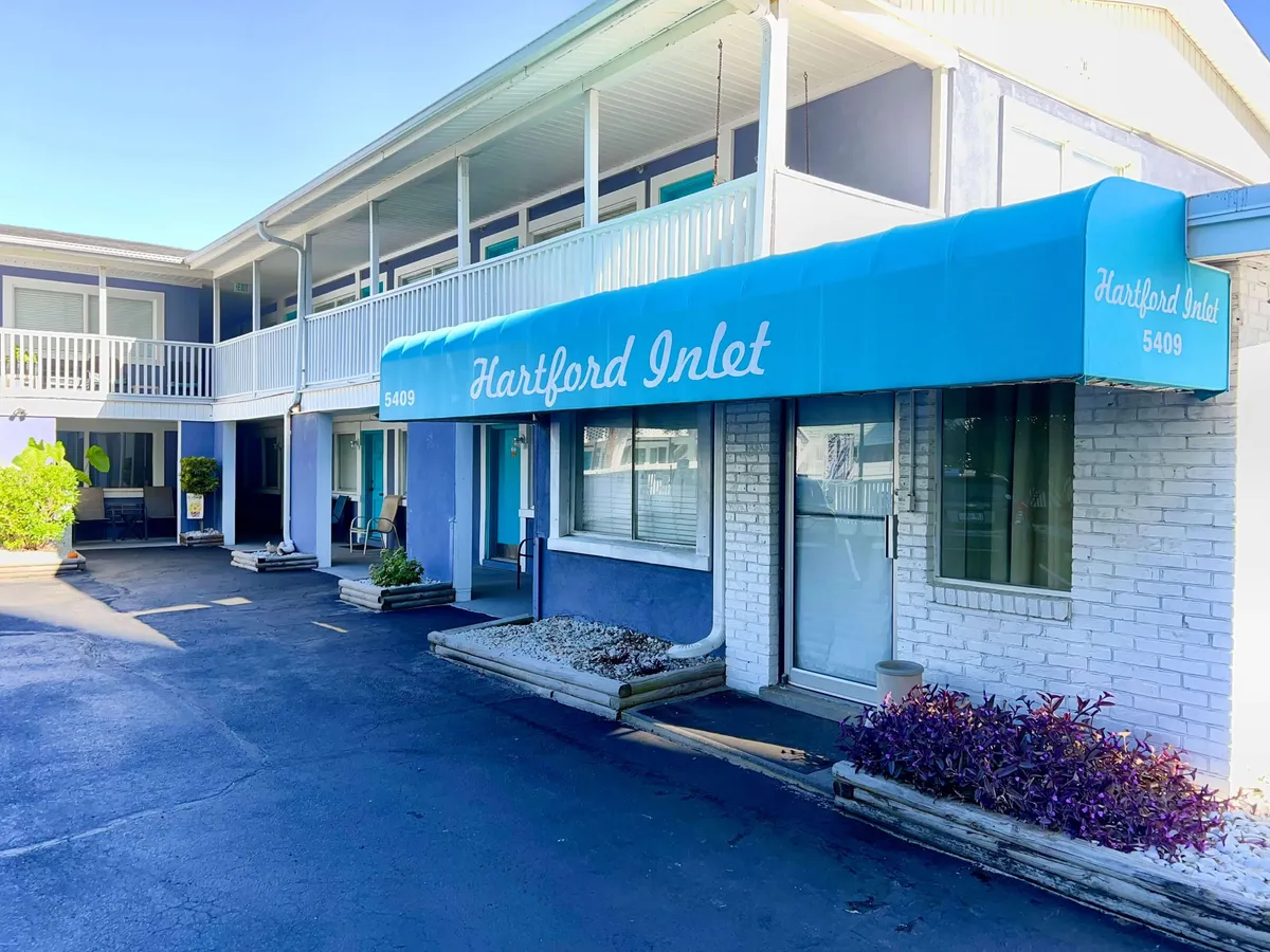 The welcoming exterior of the property, featuring a blue awning with the 'Hartford Inlet' sign, white brick wall, and lush green and purple decorative plants. The scene suggests easy access, a friendly first impression, and a relaxing outdoor environment.