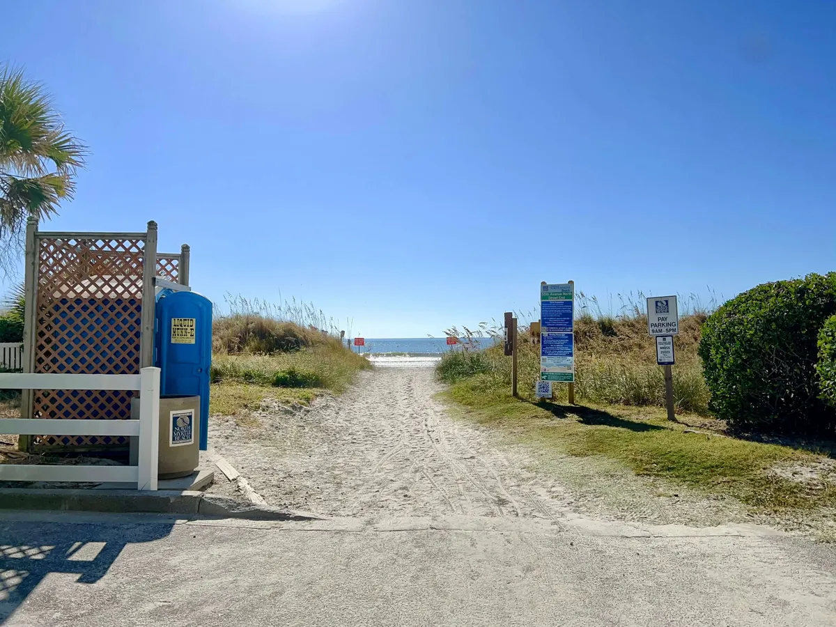 A sandy pathway leads through lush greenery directly to the beach, with information signs and a blue portable restroom visible. The inviting path hints at easy access from the condo to the shoreline, promising mornings and sunsets by the sea.
