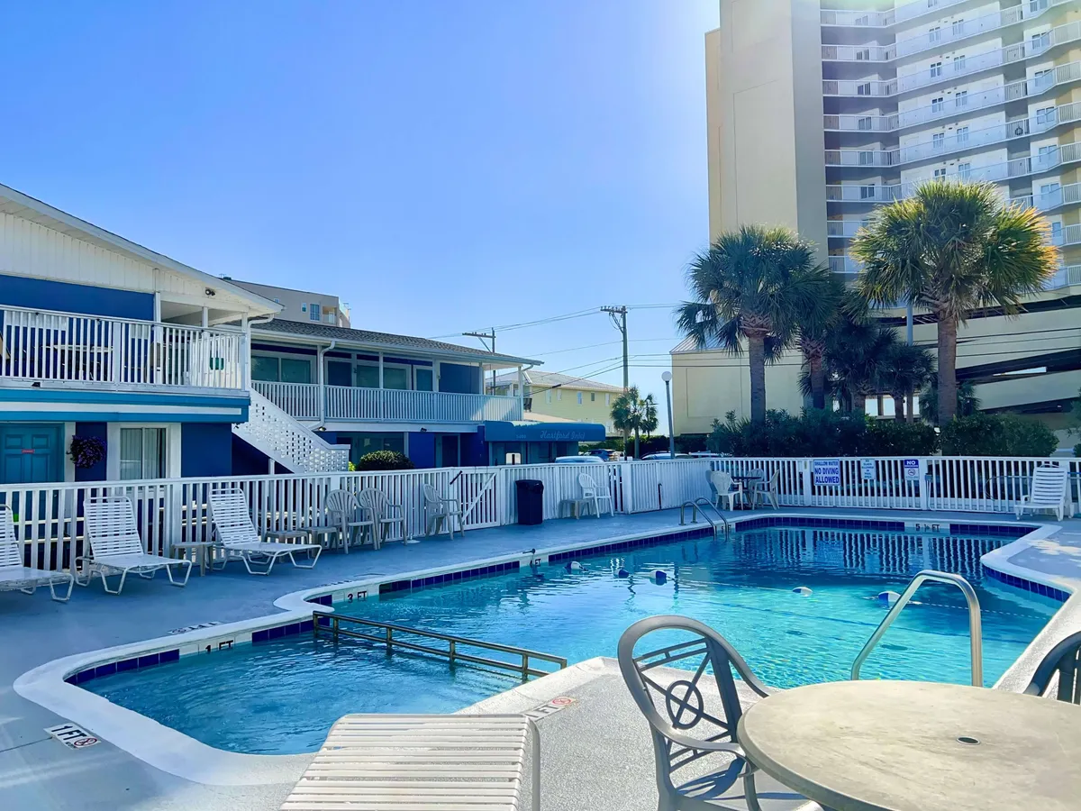 A bright, inviting pool area framed by palm trees and lounge chairs, with the building's exterior in the background. The space is ideal for relaxing, sunbathing, or socializing with friends and family, suggesting year-round enjoyment.