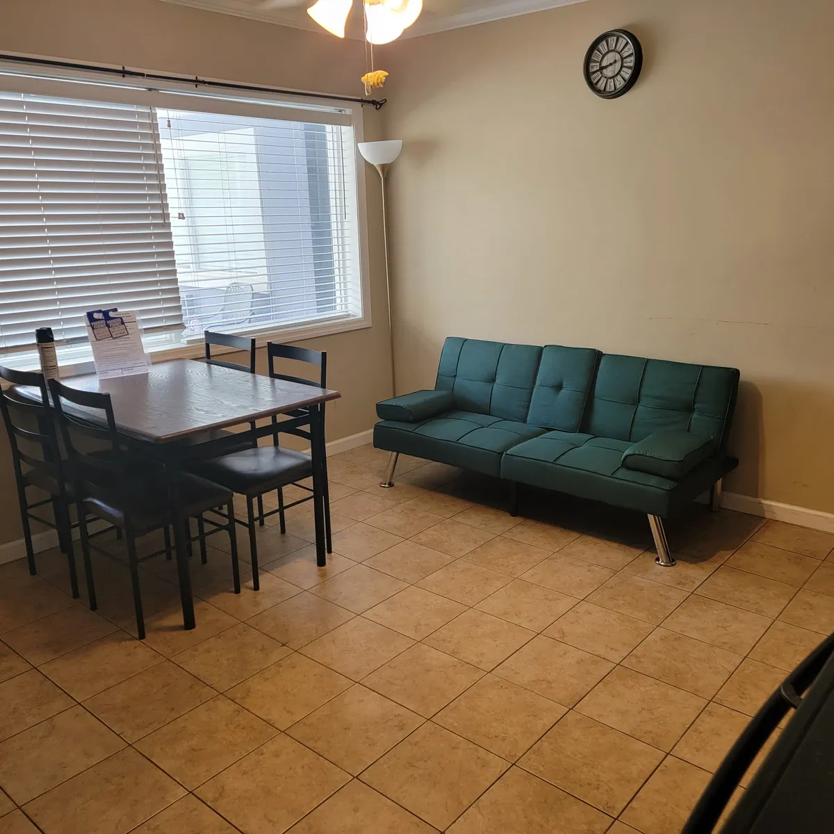 This image shows a dining table adjacent to a green sofa, with a large window letting in ample sunlight. A floor lamp and a wall clock add homely touches, and the setup invites both dining and relaxing, perfect for families and groups of friends.