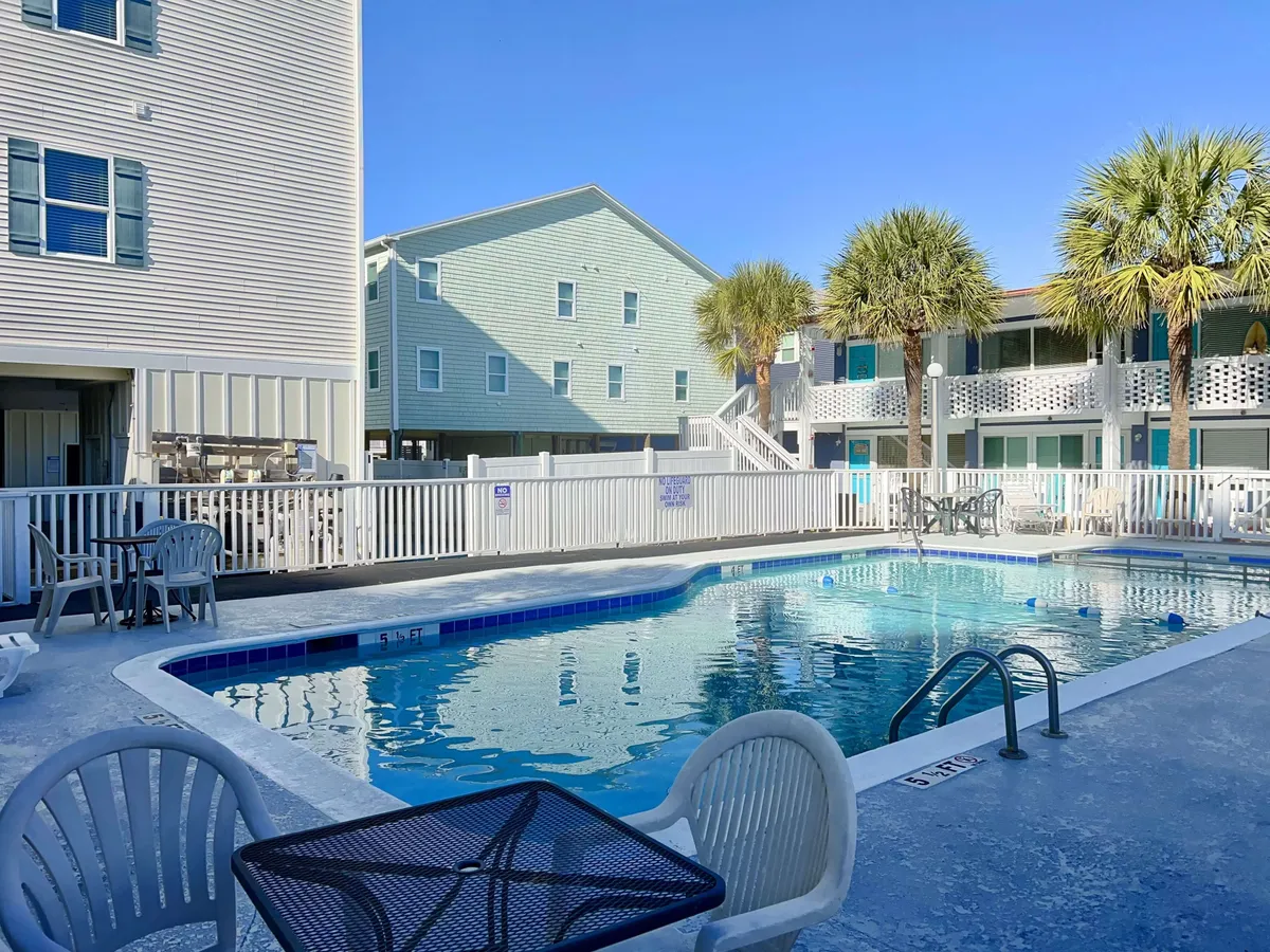 A sparkling outdoor swimming pool is the focal point, surrounded by palm trees and inviting seating areas. The clear blue water is set against a backdrop of lush greenery, providing a relaxing resort-like atmosphere. The space is designed for both activity and relaxation, with ample room for swimming, lounging, and gathering with friends or family.