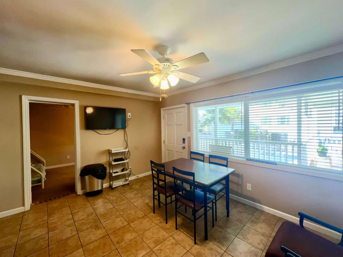 This image captures a comfortable dining area with a table and chairs, window with blinds, a ceiling fan, a television, and a doorway leading to adjoining spaces. The natural light streaming in through the window adds to the room’s inviting atmosphere, while the open layout makes it easy to imagine family meals, movie nights, or relaxed social gatherings.